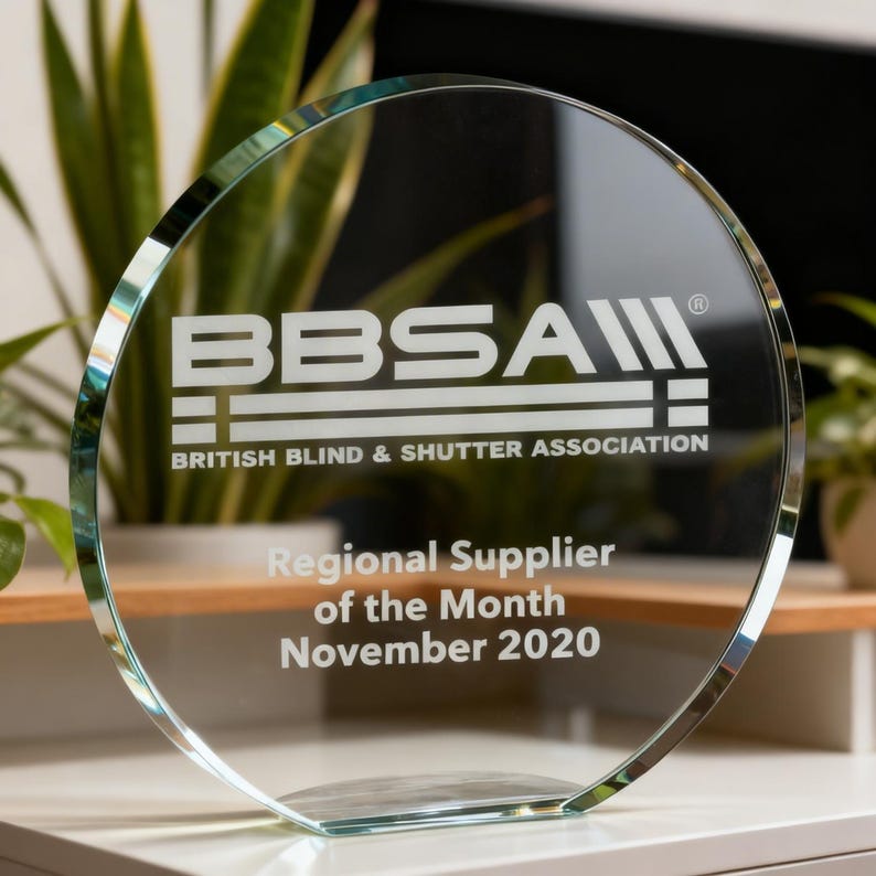 Może przedstawiać: Okrągła nagroda z przezroczystego szkła z wygrawerowanym logo BBSA British Blind & Shutter Association oraz napisem Regional Supplier of the Month November 2020. Nagroda posiada płaską podstawę umożliwiającą postawienie na powierzchni.