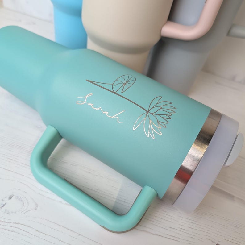May include: A teal insulated tumbler with a handle, featuring a floral design and the name "Sarah" in gold lettering. The tumbler has a silver band near the top and a white lid. Other tumblers in blue, beige, and grey are visible in the background.
