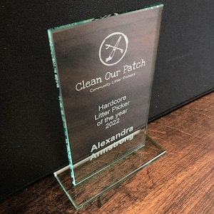 Peut inclure: Un prix en verre transparent avec une base rectangulaire et un sommet inclin&eacute; plus haut. Le texte "Clean Our Patch" et "Hardcore Litter Picker of the year 2022" est grav&eacute; sur le devant, ainsi qu'un logo et le nom "Alexandra Armstrong".