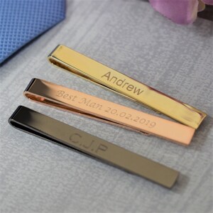 May include: Three tie clips with different engravings. The top clip is gold and engraved with the name "Andrew". The middle clip is rose gold and engraved with "Best Man 20.02.2019". The bottom clip is black and engraved with "G.J.P."