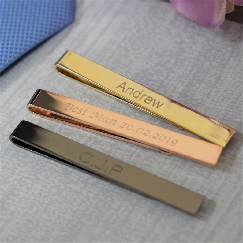 May include: Three tie clips with different engravings. The top clip is gold and engraved with the name "Andrew". The middle clip is rose gold and engraved with "Best Man 20.02.2019". The bottom clip is black and engraved with "G.J.P."