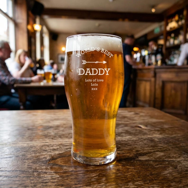 May include: A pint glass filled with golden beer and a foamy head. The glass is etched with the words "World's Best Daddy" and "Lots of love Lola xxx". The glass sits on a wooden table in a pub setting.
