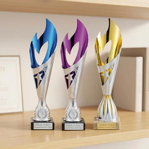 Puede incluir: Tres trofeos con la parte superior en forma de llama en azul, morado y dorado. Cada trofeo tiene un cuerpo plateado con un diseño de estrella y una base negra o beige. Las bases están etiquetadas como "Primer Lugar", "Segundo Lugar" y "Tercer Lugar" para el "Campeonato 2024".