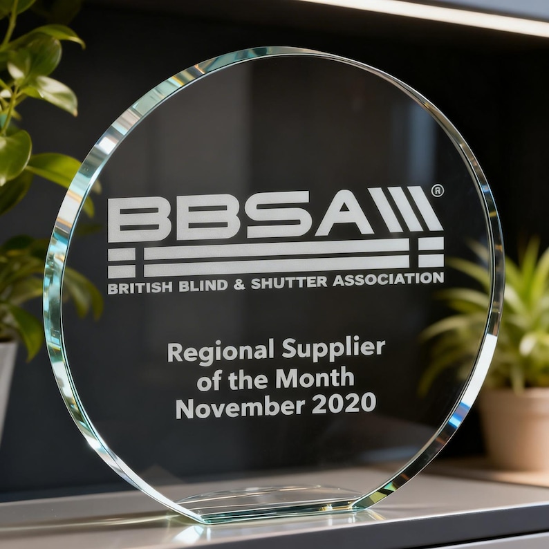 Może przedstawiać: Okrągła nagroda z przezroczystego szkła z białym grawerowanym logo BBSA British Blind & Shutter Association. Tekst głosi Regional Supplier of the Month November 2020. Nagroda posiada płaską podstawę oraz polerowaną, fazowaną krawędź.
