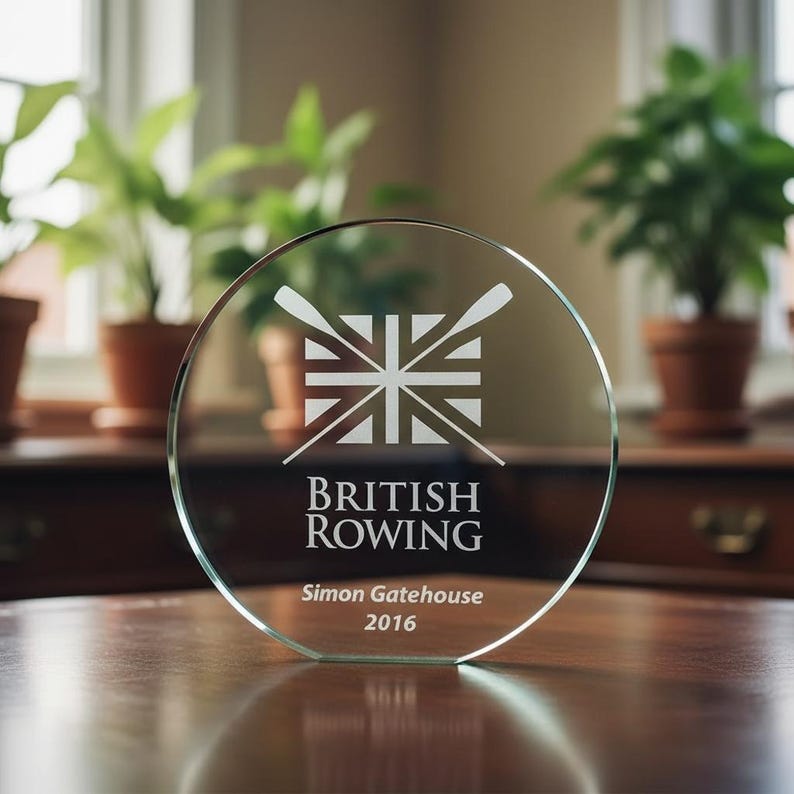 Może przedstawiać: Okrągła szklana nagroda z wygrawerowaną grafiką stylizowanej flagi brytyjskiej i dwoma skrzyżowanymi wiosłami. Tekst zawiera napis BRITISH ROWING, Simon Gatehouse oraz 2016. Nagroda ma płaską podstawę i stoi na gładkiej powierzchni.