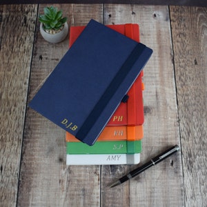 May include: A stack of personalized notebooks in various colors, including navy blue, red, orange, green, and white. Each notebook has a gold-colored initial or name. A black pen rests on a wooden surface next to the notebooks.