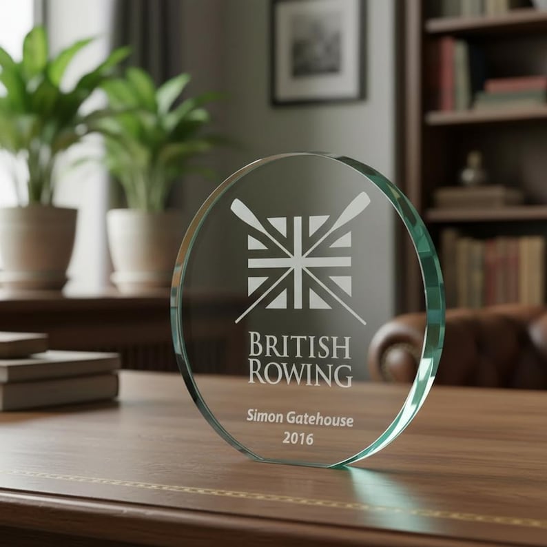 Może przedstawiać: Okrągła szklana nagroda z wygrawerowanym wzorem dw&oacute;ch skrzyżowanych wioseł na tle flagi Union Jack. Tekst zawiera napis British Rowing, Simon Gatehouse oraz 2016. Przezroczysta szklana tabliczka posiada płaską podstawę zapewniającą stabilność.
