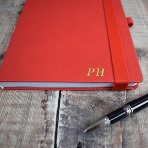 May include: A red notebook with a matching elastic band closure and gold-colored initials "PH". A black and silver pen rests on a wooden surface next to the notebook.