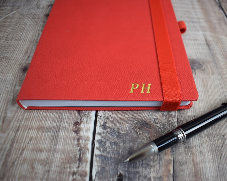 May include: A red notebook with a matching elastic band closure and gold-colored initials "PH". A black and silver pen rests on a wooden surface next to the notebook.