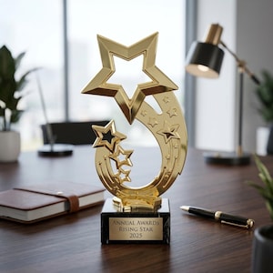 Puede incluir: Un trofeo dorado con una gran estrella en la parte superior y estrellas más pequeñas debajo. Se encuentra sobre una base de mármol negro con el texto "ANNUAL AWARDS RISING STAR 2025". El trofeo está sobre un escritorio de madera.