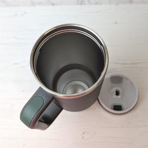 May include: A dark grey travel mug with a stainless steel rim and a black handle with a green accent. The lid is off to the side. The mug is designed to keep beverages hot or cold.