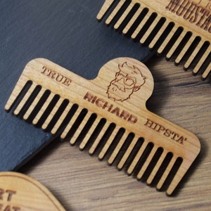 Peut inclure: Peigne &agrave; barbe en bois avec le texte "TRUE RICHARD HIPSTA" et une illustration de dessin anim&eacute; d'un homme avec une barbe et des lunettes. Le peigne est brun clair avec des dents espac&eacute;es uniform&eacute;ment.