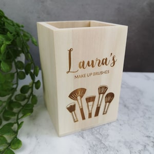 May include: A light wood makeup brush holder with the text "Laura's Make Up Brushes" and an engraved design of makeup brushes.