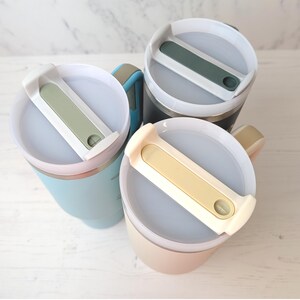 May include: Three insulated travel mugs in pastel shades: light blue, beige, and dark grey. Each mug features a white lid with a coloured slider and a matching handle. The mugs are designed to keep drinks hot or cold.