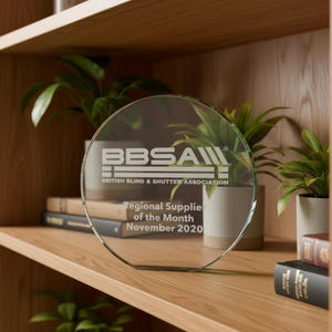 Może przedstawiać: Okrągła szklana tabliczka z wygrawerowanym logo BBSA oraz napisem British Blind & Shutter Association, Regional Supplier of the Month, November 2020. Trofeum z przezroczystego szkła ma płaską podstawę i stoi na drewnianej p&oacute;łce.