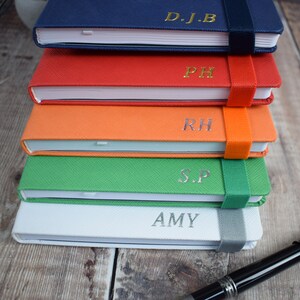 May include: A stack of five personalized notebooks in various colors: blue, red, orange, green, and white. Each notebook has a matching elastic band closure and is embossed with initials or a name in gold or silver. A black pen rests on the wooden surface.