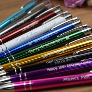 Puede incluir: Primer plano de una variedad de bol&iacute;grafos personalizados con cuerpos de metal de diferentes colores y mensajes grabados. Los bol&iacute;grafos est&aacute;n dispuestos en fila, con los mensajes grabados mirando al espectador. Los mensajes incluyen nombres, fechas y ocasiones especiales.
