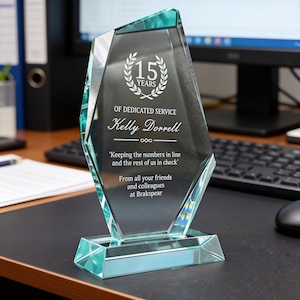 May include: A clear, faceted glass award with a teal base. The award is engraved with "15 YEARS", "OF DEDICATED SERVICE", a name, and a quote. The text is in white. The award is displayed on a black desk.