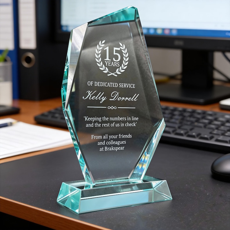 May include: A clear, faceted glass award with a teal base. The award is engraved with "15 YEARS", "OF DEDICATED SERVICE", a name, and a quote. The text is in white. The award is displayed on a black desk.