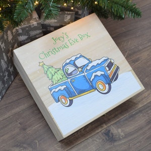 Peut inclure: Boîte de Noël en bois clair avec une illustration d'un camion bleu transportant un sapin de Noël. La boîte porte l'inscription "Joey's Christmas Eve Box" en vert. De la neige est représentée sur le camion et le sol. La boîte est sur une surface en bois.
