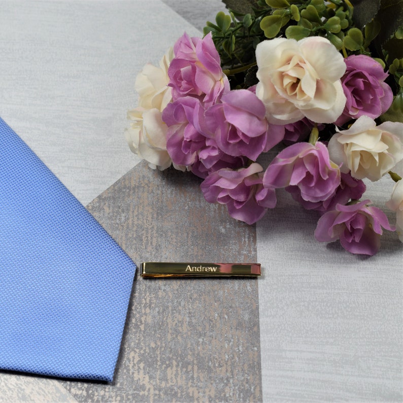 May include: A gold tie bar with the name "Andrew" engraved on it. The tie bar is lying on a light blue patterned fabric and a white surface with a bouquet of pink and white roses.