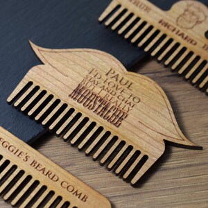 Peut inclure: Peigne &agrave; moustache en bois clair avec le texte "PAUL I'D LOVE TO STAY AND CHAT BUT I REALLY MOUSTACHE". Le peigne est en forme de moustache. D'autres peignes sont visibles avec du texte.