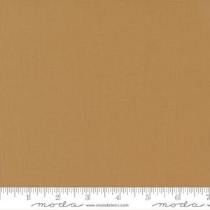 May include: A close-up of a solid, tan-colored fabric with a subtle woven texture. A ruler is placed at the bottom of the fabric, with measurements in inches. The fabric appears to be a quilting cotton.