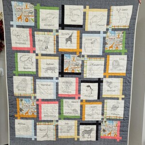 May include: A handmade quilt featuring a gray background with a grid of squares. Each square displays a different animal illustration with its name, including a giraffe, zebra, and penguin. The quilt is bordered with a light blue and pink trim.