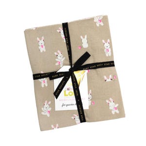 May include: A stack of folded fabric in a light tan color featuring a repeating pattern of white cartoon rabbits wearing pink headphones and holding pink devices. The fabric is tied with a black ribbon printed with the text Ruby Star Society.