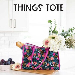 May include: A dark blue fabric tote bag featuring a vibrant floral pattern with pink, orange, and green flowers, accented by bright pink trim and handles. The bag contains a baguette and a bouquet of white flowers, with text reading ALL THE THINGS TOTE.