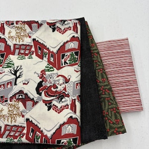 May include: A stack of Christmas-themed fabric pieces. One fabric features a Santa Claus design with red houses and snow. Other fabrics include a black textured fabric, a green fabric with red berries, and a red and white striped fabric.