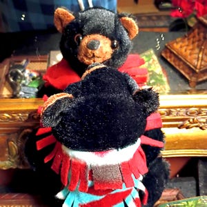 May include: Two plush black bears with brown snouts and ears. One bear is in the foreground, its back to the viewer, wearing a red and turquoise fringed garment. The other bear is in the background, wearing a red scarf.