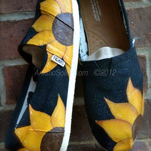 May include: A pair of black canvas shoes with white trim, painted with yellow sunflowers. The shoes have a brown leather insole with the text "TOMS" printed on it. The text "With every pair you purchase, TOMS will give a pair of new shoes to a child in need. One for One" is printed on the insole.