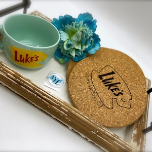 May include: A wooden tray with a light blue mug that says "Luke's" on it. There are two cork coasters with the same logo on them. The coasters say "Luke's" and "Stars Hollow, CT".