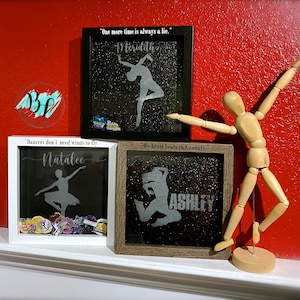 May include: Three shadow box frames with different colored backgrounds and glitter. Each frame has a dancer silhouette and a personalized name. The frames are on a white shelf with a wooden mannequin in the background.