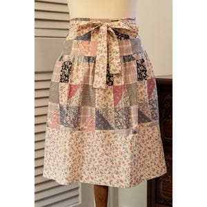 May include: A patchwork apron with a floral print bottom and a patterned top. The apron features a tie-waist design and is displayed on a mannequin. The patchwork includes pink, black, and gray squares and triangles.