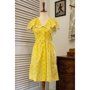 May include: A vintage yellow dress with a floral pattern and white lace trim. The dress features a V-neck, short sleeves, and a button-down front. The dress is displayed on a mannequin.