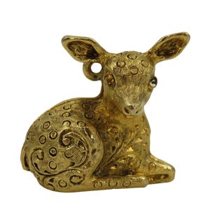 May include: A gold-toned metal charm shaped like a resting fawn. The surface features a textured, engraved pattern of circles and swirls across the body and head. A small circular loop is attached to the back of the head for hanging purposes.