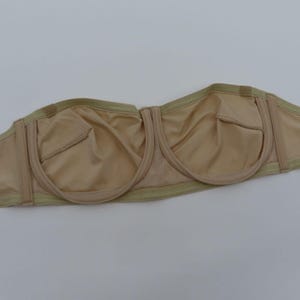May include: A beige strapless bra with a smooth, seamless design. The bra features molded cups and a supportive underwire. The edges are trimmed with a light green fabric. The bra is designed for a comfortable and secure fit.
