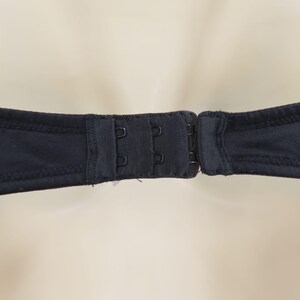 Op de afbeelding: Close-up van een zwarte bh-sluiting met drie haak-en-oog sluitingen. De stof van de bh is effen zwart en de sluiting is gecentreerd in het beeld. De achtergrond is een zachte, neutrale kleur.