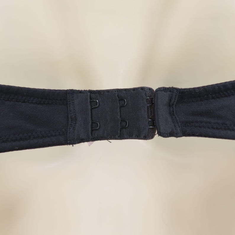 Op de afbeelding: Close-up van een zwarte bh-sluiting met drie haak-en-oog sluitingen. De stof van de bh is effen zwart en de sluiting is gecentreerd in het beeld. De achtergrond is een zachte, neutrale kleur.