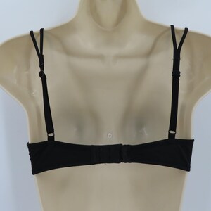 Op de afbeelding: Zwarte beha met verstelbare bandjes en een haak-en-oogsluiting. De beha wordt getoond op een mannequin, waarbij het rugontwerp te zien is. De bandjes zijn dun en de stof lijkt glad.