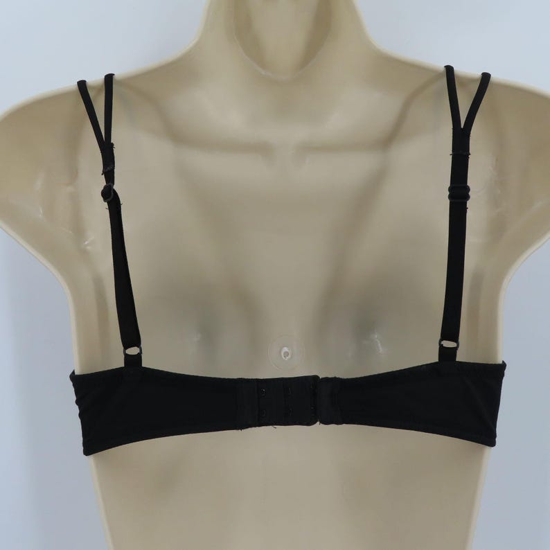 Op de afbeelding: Zwarte beha met verstelbare bandjes en een haak-en-oogsluiting. De beha wordt getoond op een mannequin, waarbij het rugontwerp te zien is. De bandjes zijn dun en de stof lijkt glad.