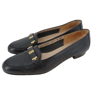 Puede incluir: Un par de mocasines de cuero negro con un acabado texturizado. Cada zapato presenta una hebilla dorada en el empeine. Los zapatos tienen un diseño clásico sin cordones y un tacón bajo.