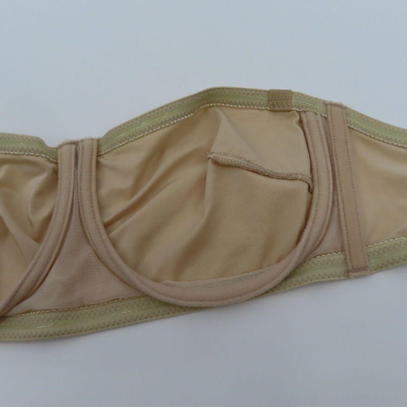 May include: A beige strapless bra with a smooth, silky texture. The bra features a structured design with underwire support and adjustable closures. The edges are trimmed with a delicate, light green stitching.