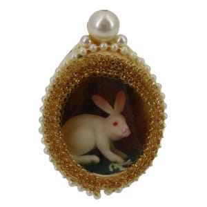 May include: An egg-shaped ornament with a white rabbit inside, framed by gold trim and small pearls. A large pearl sits at the top. The rabbit has red eyes. The background is a dark brown color.