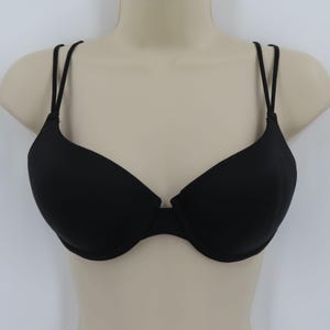 Op de afbeelding: Zwarte beha met dunne, dubbele bandjes. De beha wordt getoond op een mannequin buste tegen een witte achtergrond. De beha heeft een klassiek ontwerp met voorgevormde cups en een middenstuk.