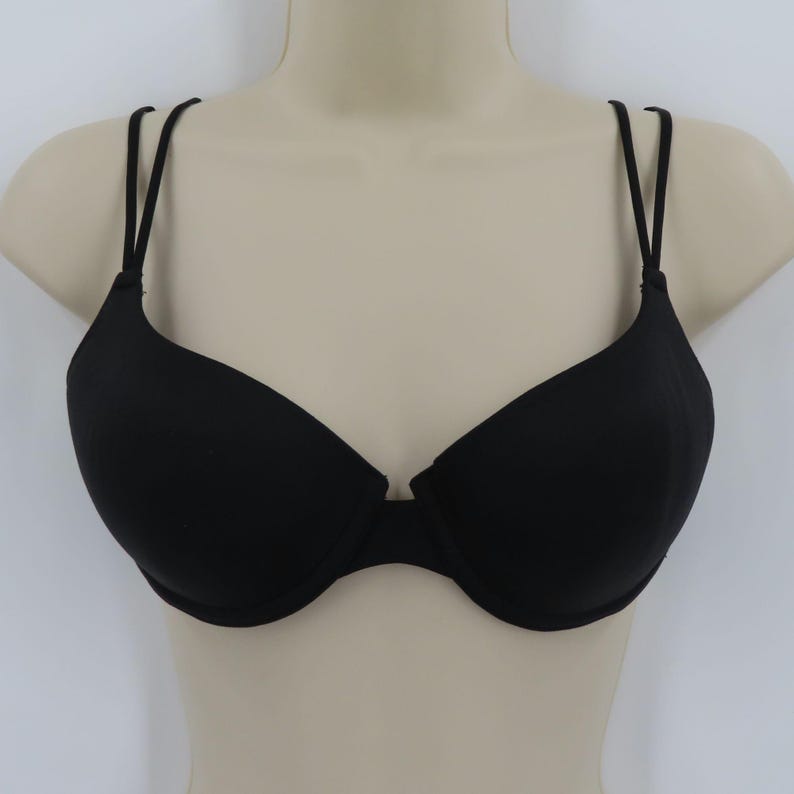 Op de afbeelding: Zwarte beha met dunne, dubbele bandjes. De beha wordt getoond op een mannequin buste tegen een witte achtergrond. De beha heeft een klassiek ontwerp met voorgevormde cups en een middenstuk.