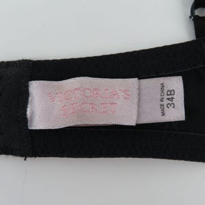 Op de afbeelding: Close-up van een zwarte bh met een label met de tekst "VICTORIA'S SECRET" in roze letters. Het label geeft ook de maat 34B en "MADE IN CHINA" aan. De bh is gemaakt van een glad, waarschijnlijk synthetisch materiaal.