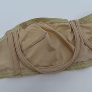 May include: A beige strapless bra with a smooth, seamless design. The bra features a structured underwire and a soft, neutral fabric. The interior lining is a matching beige color, and the edges are trimmed with a delicate, light green stitching.